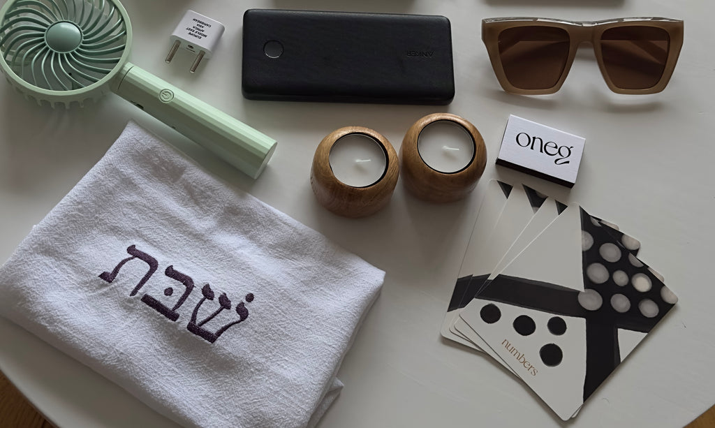 A flatlay of travel items including Oneg's Shabbat ritual items, travel items like a portable fan and charger, sunglasses, a reading tablet, a journal, and a pair of headphones.